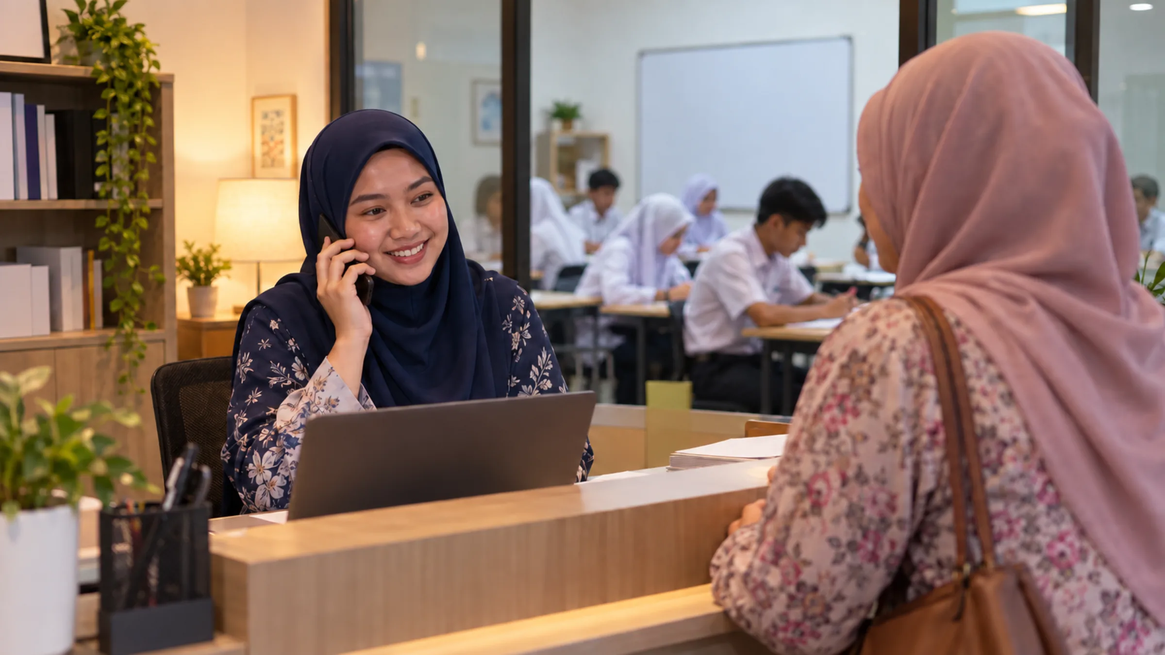 An administrator in a navy tudung answers a phone at a tuition centre with students in the classroom behind her.