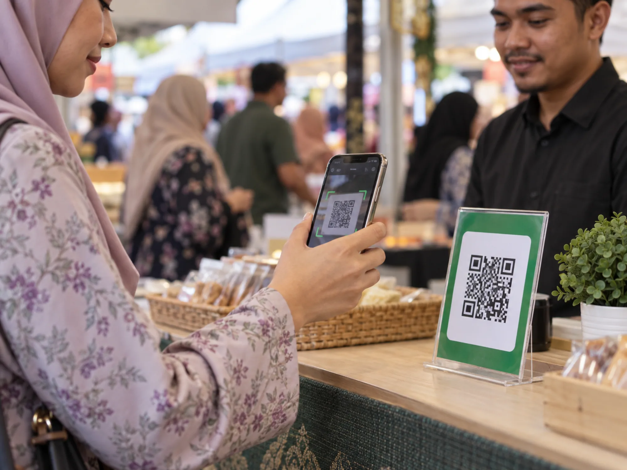 A hand in a long sleeve holds a phone toward a QR code stand with a green border at an event counter.